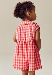 Bambino piccolo con capelli ricci raccolti, che indossa un vestito smanicato a quadretti rossi e bianchi, in piedi di fronte a una parete beige chiaro.