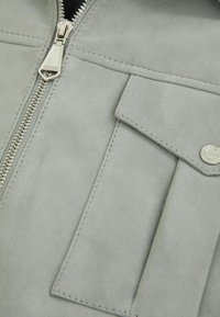 Jachetă din suede gri, cu fermoar metalic, un buzunar cu clapetă și detalii cusute, evidențiind o textura netedă și un design minimalist.