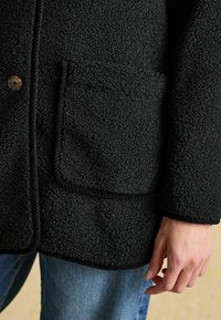 Musta tekstuuri takki pörröise pinnaga. Omabnaastu taskut, köisdetaili ja nööpkatet. Kantud siniste teksadega.