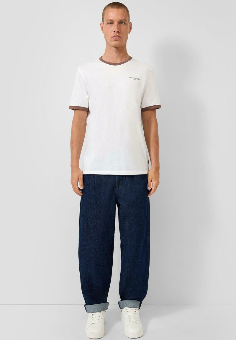 Mladý muž stojící rovně, oblečený v bílém tričku s hnědým lemováním a tmavě modrých zkrácených džínách s bílými teniskami, na pozadí obyčejné stěny.