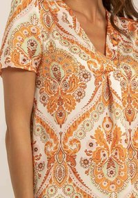 Haut à manches courtes avec un motif floral orange et vert clair sur un fond pâle. Encolure en V avec un détail froncé au centre.