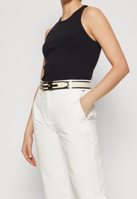 Canottiera a coste blu navale abbinata a pantaloni bianchi a vita alta con una cintura nera e crema. Mano visibile nella tasca del pantalone.