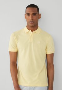 Světle žluté polo tričko s límečkem, krátkými rukávy a malým logem na hrudi. Měkká textura, volný střih a minimalistický design.