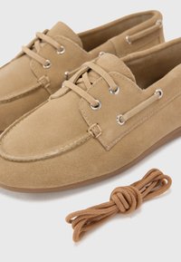 Beige Wildleder Slipper mit runder Spitze, zwei Ösen-Design und kontrastierenden braunen Schnürsenkeln. Enthält eine zusätzliche passende Schnur, die daneben gelegt ist.