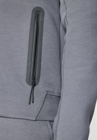 Grå sweatshirt med en lynlåslomme på venstre arm, med en glat stofstruktur og kontrasterende sort lynlåsdetalje.