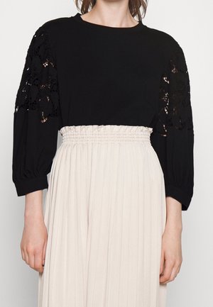 Blusa negra con recortes de encaje floral en las mangas abullonadas de tres cuartos, combinada con falda plisada beige de talle alto con cinturilla fruncida.