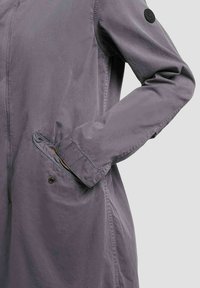 Gros plan d'une veste mauve à manches longues avec une main dans la poche avant et un patch circulaire noir sur la partie supérieure de la manche.