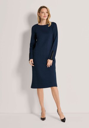 Vrouw draagt een marineblauwe midi jurk met lange mouwen en knoopdetails op de mouwen, en zwarte hoge hakken, staand tegen een effen achtergrond.