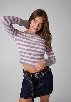 Ung kvinde med langt brunt hår iført en lyserød og grå stribet cropped sweater og mørk denim nederdel med sort bælte og tørklæde.