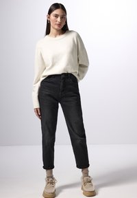Kremowy sweter wełniany, czarne spodnie typu high-waist, beżowe puszyste buty i jasnobrązowe skarpetki widoczne. Prosty design, luźny krój.