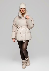 Béžová puffer bunda s vysokým límcem, páskovým pasem a prošívanou texturou, v kombinaci s lesklými hnědými legínami a béžovými botami. Neutrální čepice.