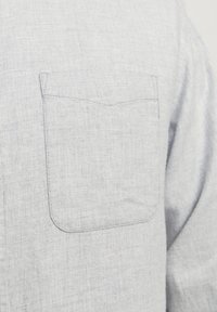 Chemise en tissu gris clair texturé avec une poche poitrine unique et des coutures visibles à l’avant.