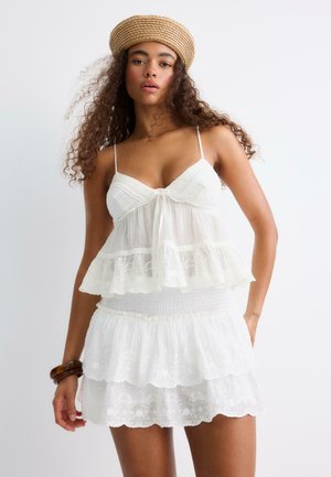 Femme aux cheveux bouclés portant un chapeau tissé beige, une robe blanche en dentelle à volants avec fines bretelles, et un bracelet foncé au poignet, sur un fond uni.