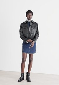 Veste cropped en cuir noir avec des boutons sur le devant et deux poches poitrine, associée à une mini-jupe en denim et des bottines noires.