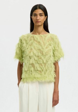 Kvinde iført en limegrøn tekstureret kortærmet top med frynser og højtaljede hvide plisserede bukser.