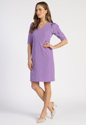 Lavendelfarbenes Kleid mit V-Ausschnitt und kurzen Ärmeln, tailliert mit geradem Rock. Trägt beigen Schuhe mit Schleifenakzenten.