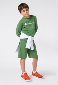 Jongen in groene Champion sweatshirt en shorts met een witte sweatshirt om zijn middel geknoopt, met oranje sneakers en witte Champion sokken.