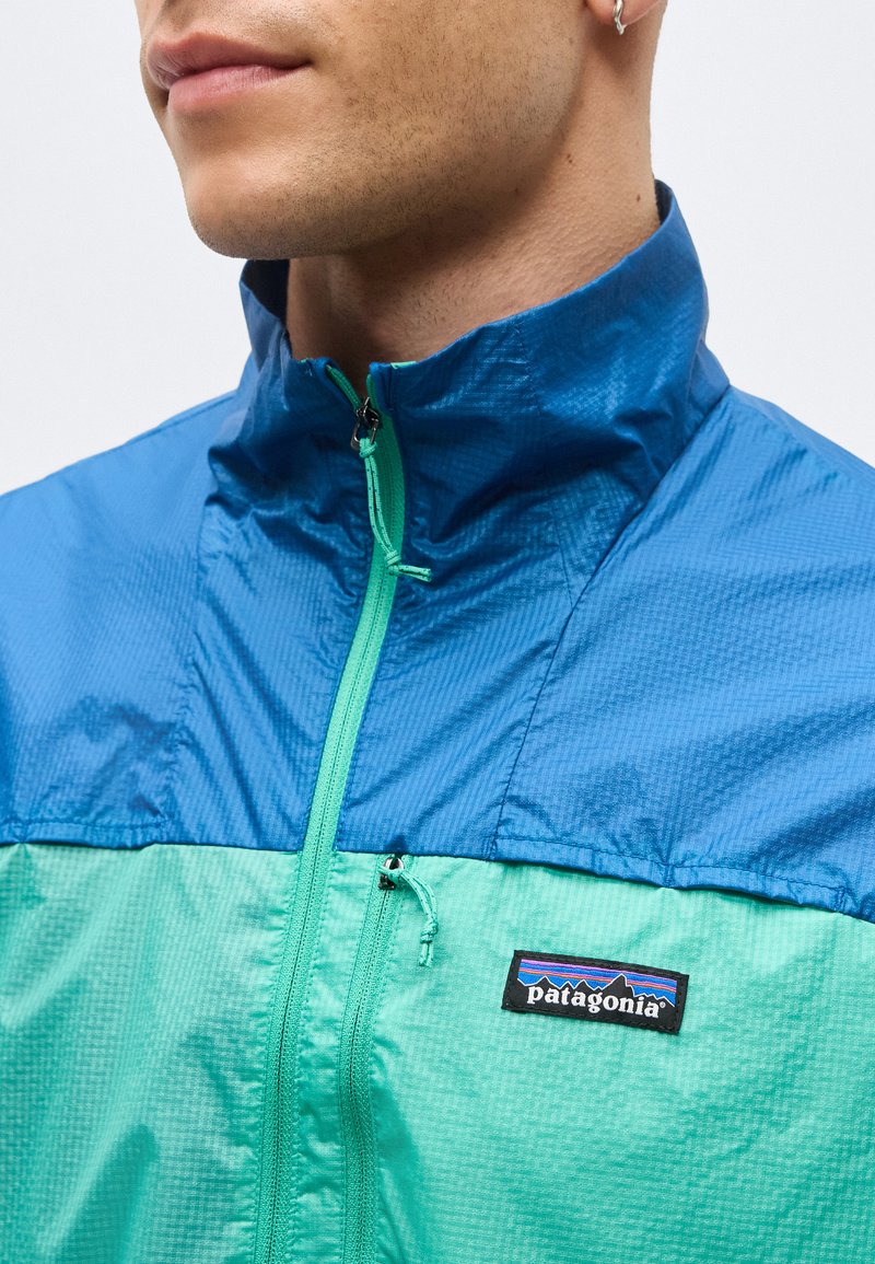 Mann trägt eine blaue und türkisfarbene Patagonia-Jacke mit hohem Kragen und sichtbarem Patagonia-Logo auf der Brust.