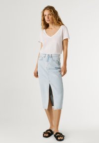 Donna in piedi che indossa una maglietta bianca con scollo a V, gonna midi in denim celeste a vita alta con spacco frontale e sandali neri borchiati.