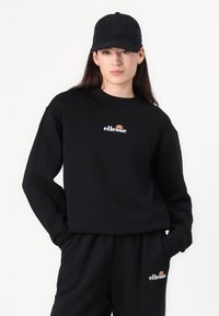Svart hettegenser og bukser med "ellesse"-logo. Materialet ser mykt ut, med avslappet passform og rund hals. Også iført svart caps.