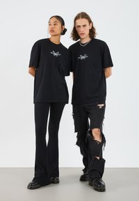 Two individuals stand side by side, wearing black graphic t-shirts, black ribbed flared pants, and distressed black jeans with rips.
