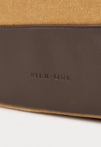 Tan canvas fabric with a brown faux leather bottom; features embossed "PIER ONE" branding. Seam shows quality stitching and texture contrast.