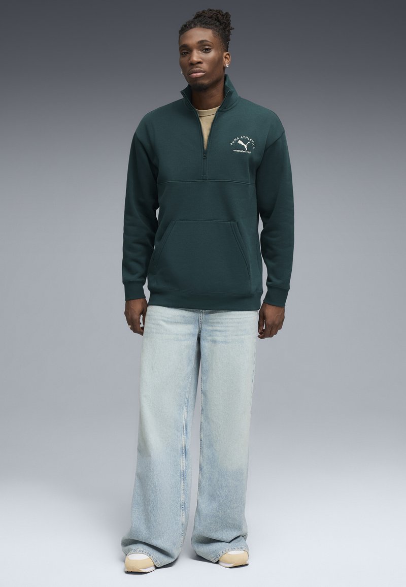 Donkergroene pullover sweatshirt met een rits-kraag, voorzien van een voorzak en een wit logo. Draagt samen met lichtblauwe wijde jeans en beige schoenen.