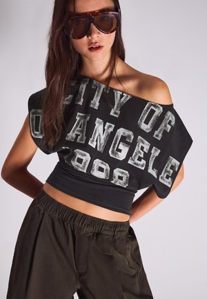 Fiatal nő, aki túlméretezett teknőcpáncél mintás napszemüveget, fekete váll nélküli crop topot visel, amin az szerepel: „City of Angeles 2000”, illetve laza sötét nadrágot.