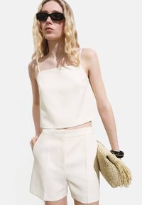 Top corto bianco con spalline sottili, abbinato a pantaloni corti coordinati. La modella tiene una borsa a tracolla beige intrecciata con frange. Lo sfondo è bianco uniforme.