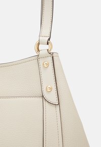Beige leren handtasriem met textuur, bevestigd met gouden ringen en gouden studs, met gedetailleerde stiknaad op een effen achtergrond.