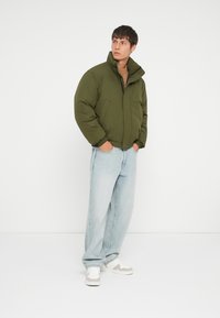Olive green puffer jacket with a high collar, worn over a beige knit sweater, paired with light blue wide-leg jeans and white sneakers.