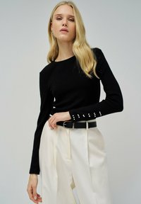 Zwarte ribbelige top met lange mouwen en een ronde halslijn, voorzien van parelaccenten op de mouwmanchetten, gecombineerd met crèmekleurige high-waisted broeken en een zwarte riem.