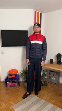 Veste matelassée marine, rouge et blanche avec une fermeture éclair complète. Pantalon large en denim bleu et chaussures marron. Cadre intérieur avec des plantes et une télévision.
