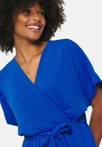 Kráľovsky modré zavinovacie šaty s krátkymi rukávmi, hlbokým výstrihom do V a opaskom na viazanie v páse. Tkanina má hladkú textúru a jemný lesk.