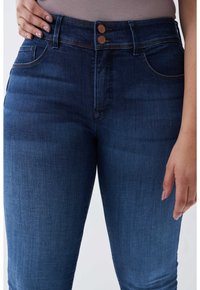 Primer plano de una persona que lleva vaqueros de talle alto azul oscuro con cierre de doble botón y bolsillos delanteros, con la mano apoyada en la cadera.