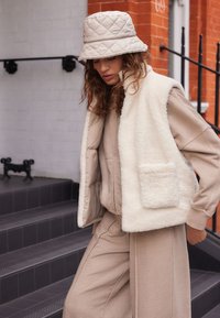 Gilet in pelliccia sintetica di colore crema sopra una felpa beige con cappuccio e pantaloni abbinati, con una tasca frontale; un cappello a secchiello beige trapuntato completa il look.