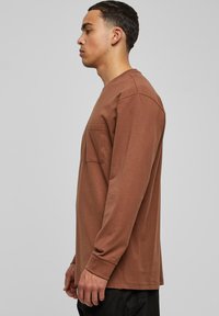 Camisa de manga larga marrón de algodón con un corte cómodo, que incluye un bolsillo en el pecho y puños de canalé, con una textura suave y un diseño sencillo.