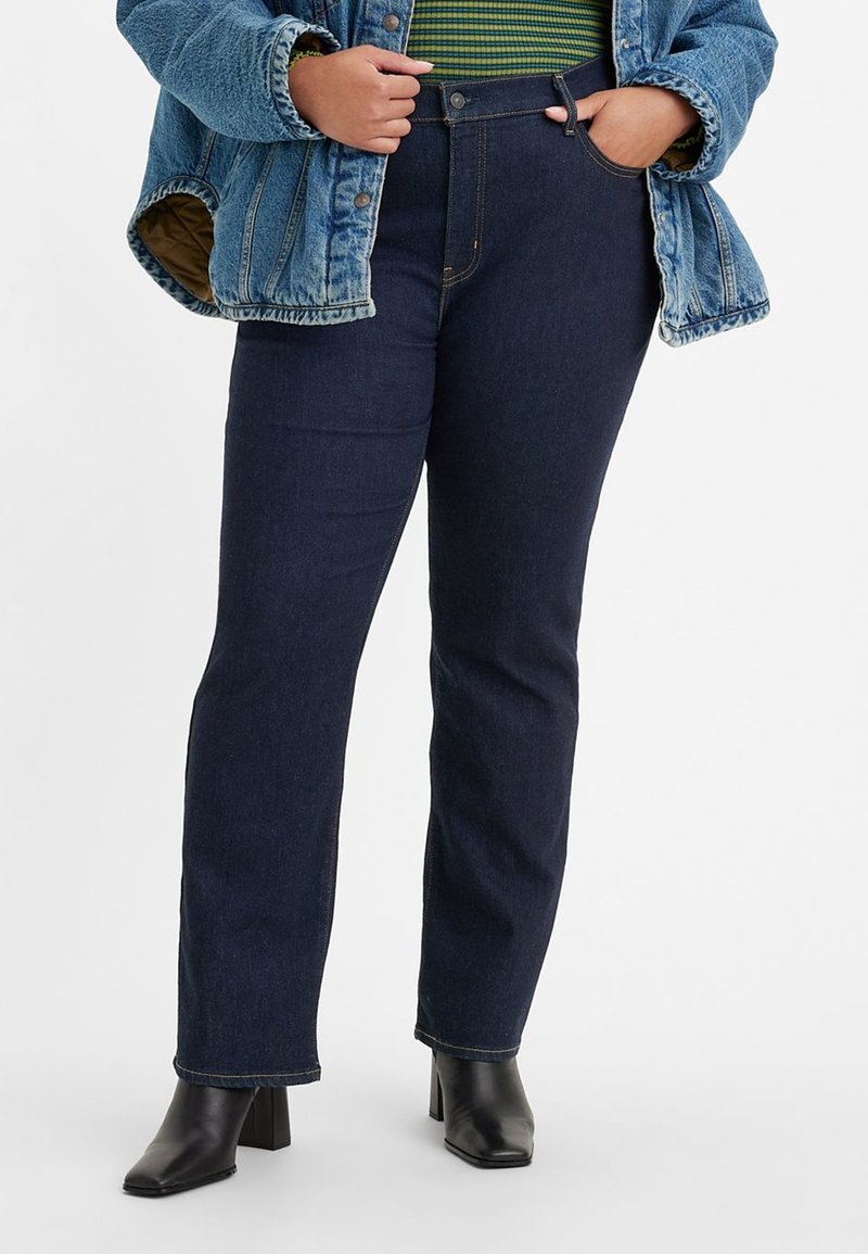 Jean en denim bleu foncé, coupe droite, taille haute et coutures contrastées, associé à une veste en denim et des bottes noires à talons.