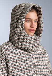 Pufasti kaput s kvadratnim uzorkom i velikim kapuljačom, s multikolor uzorkom houndstooth u boji krem, burgundac i mornarsko plava. Glatka tekstura.