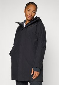 Osoba u crnoj jakni s kapuljačom stoji nasuprot jednostavne pozadine, pokazujući samouvjeren izraz lica.