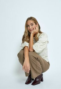 Cream blouse with puffed sleeves, brown wide-leg trousers, and burgundy high-heel boots. Simple backdrop, subject in a squatting pose.