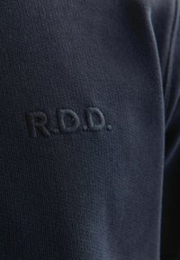Jūras zilas krāsas audums ar teksturētu dziju un izšūtu logo "R.D.D." atbilstošā diega krāsā. Auduma tuvplāns.