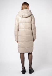 Beige pufferjas met een capuchon, met een gewatteerde textuur, lange mouwen en een rechte snit die net boven de knieën valt. Zwarte schoenen.