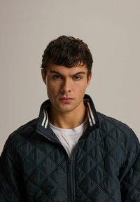 Czarna kurtka pikowana z wysokim kołnierzem i białymi paskami. Posiada zamek błyskawiczny oraz teksturowany wzór w romby na całej powierzchni.