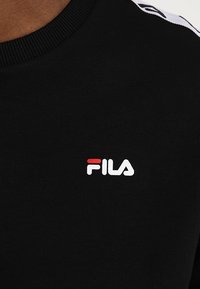 Sweat-shirt en coton noir avec un col côtelé et un petit logo Fila blanc et rouge sur la poitrine. Texture lisse avec un design minimaliste.