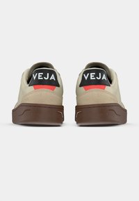 Par de zapatillas beige con suelas marrones, lengüetas negras en el talón, detalles rojos y texto blanco de la marca VEJA en la parte trasera.
