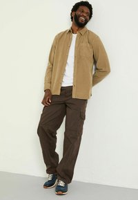 Beige Cordshirt über einem weißen T-Shirt, kombiniert mit dunkelbraunen Cargo-Hosen und blauen Sneakers mit weißen Schnürsenkeln und braunen Akzenten.