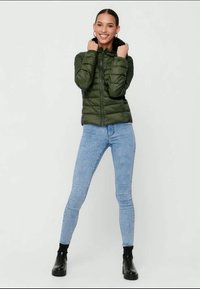 Grüne gepolsterte Jacke mit durchgehendem Reißverschluss, hohem Kragen und texturierter Oberfläche. Getragen über hellblauen Skinny-Jeans und schwarzen Ankle-Boots.