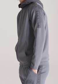 Šedý teplákový komplet s kapucí na zip, bočními kapsami a logovými akcenty na rukávech a kalhotách. Vyrobeno z hladkého, elastického materiálu.