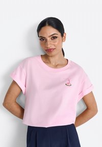 Roze katoenen T-shirt met korte opgerolde mouwen, voorzien van een klein geborduurd ontwerp op de borst. Korte lengte, zachte textuur.